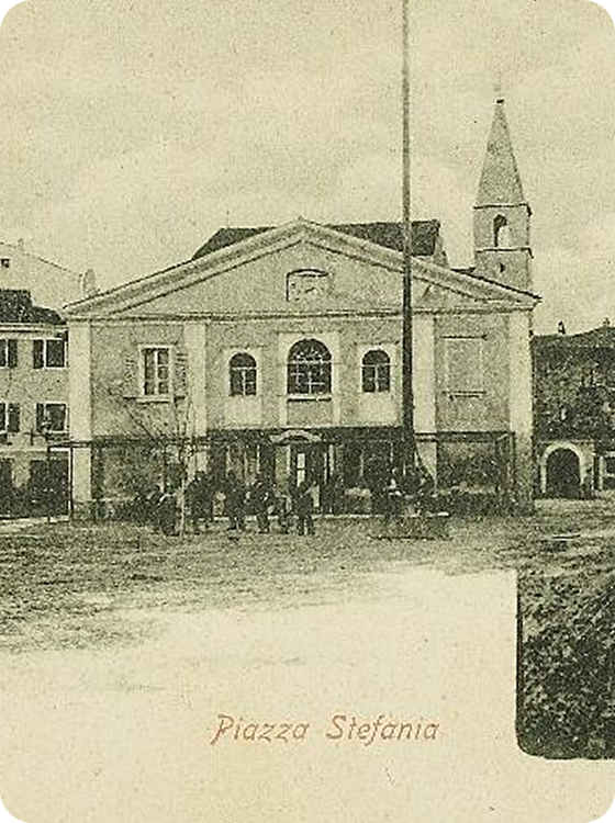 Stara sepia razglednica trga Piazza Stefania z ljudmi pred klasicistično stavbo s stebrastim pročeljem in bližnjim zvonikom, spodaj napis \"Piazza Stefania\".