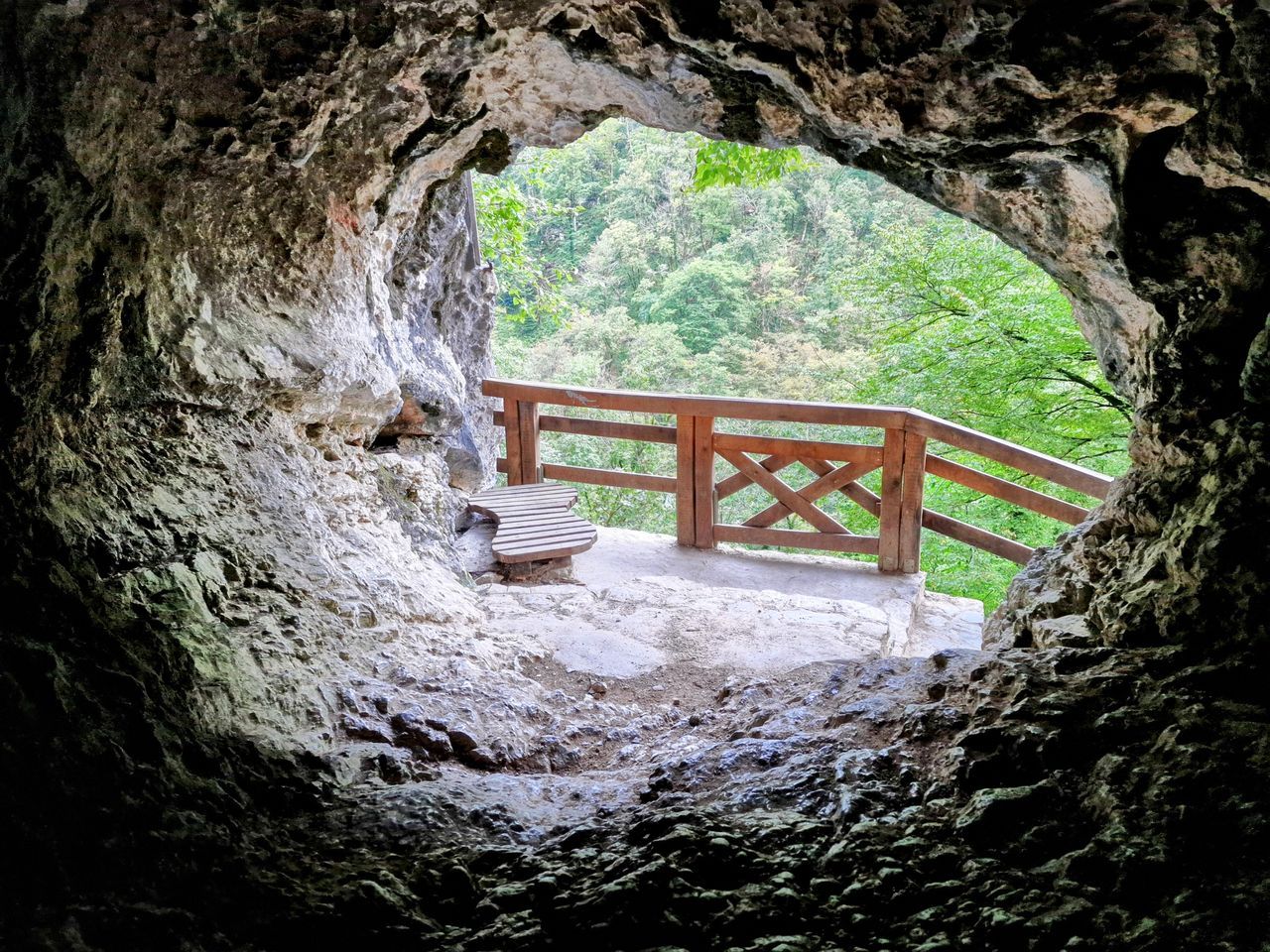 Pogled iz notranjosti jame skozi naravno odprtino na razgledno ploščad z leseno ograjo in klopjo, obdano z zelenim gozdom.