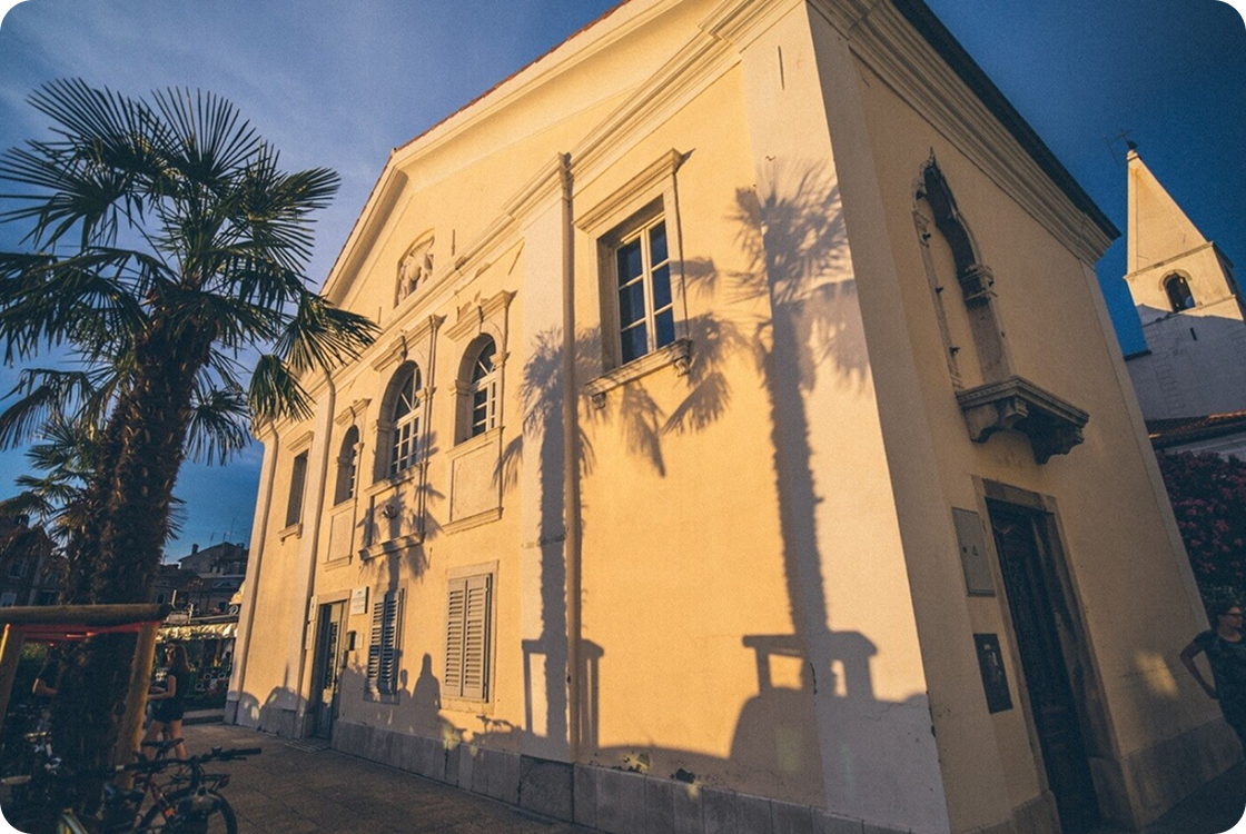 Rumeno-oker historična stavba s poudarjenimi okni in zvonikom v ozadju, ob zahajajočem soncu, ki na fasado meče dolge sence palm.